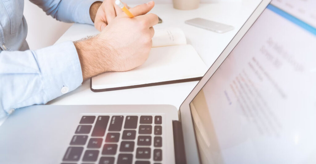 A man sits at a desk, writing in a notebook while typing on a laptop, focused on his work.
