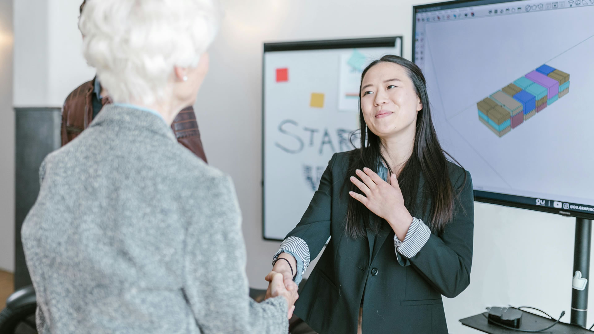 A woman shakes hands with another woman in front of a large screen displaying a presentation.
