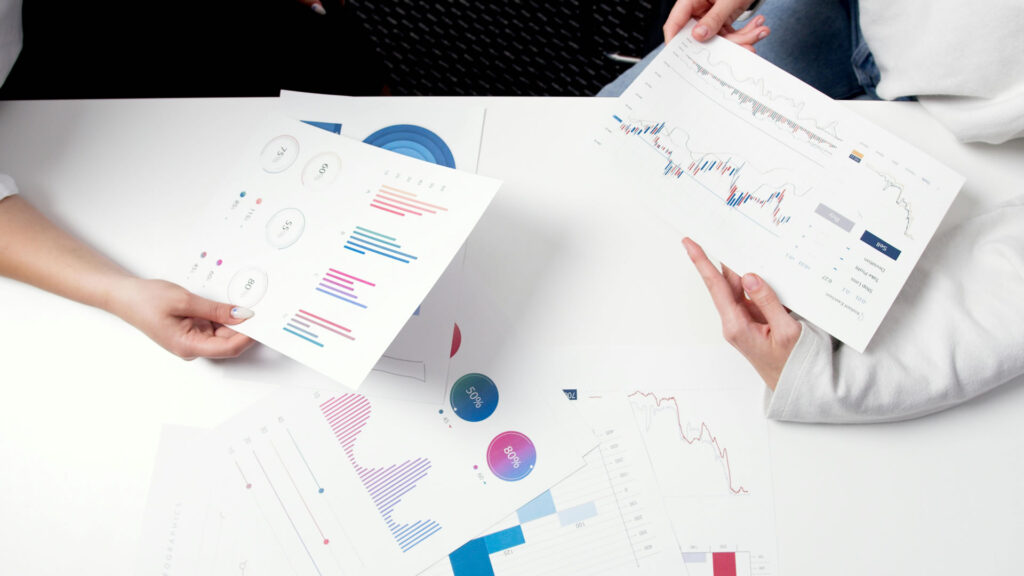 Two people analyze various charts and graphs on paper at a table, focusing on data visualizations with bar graphs, pie charts, and line graphs.