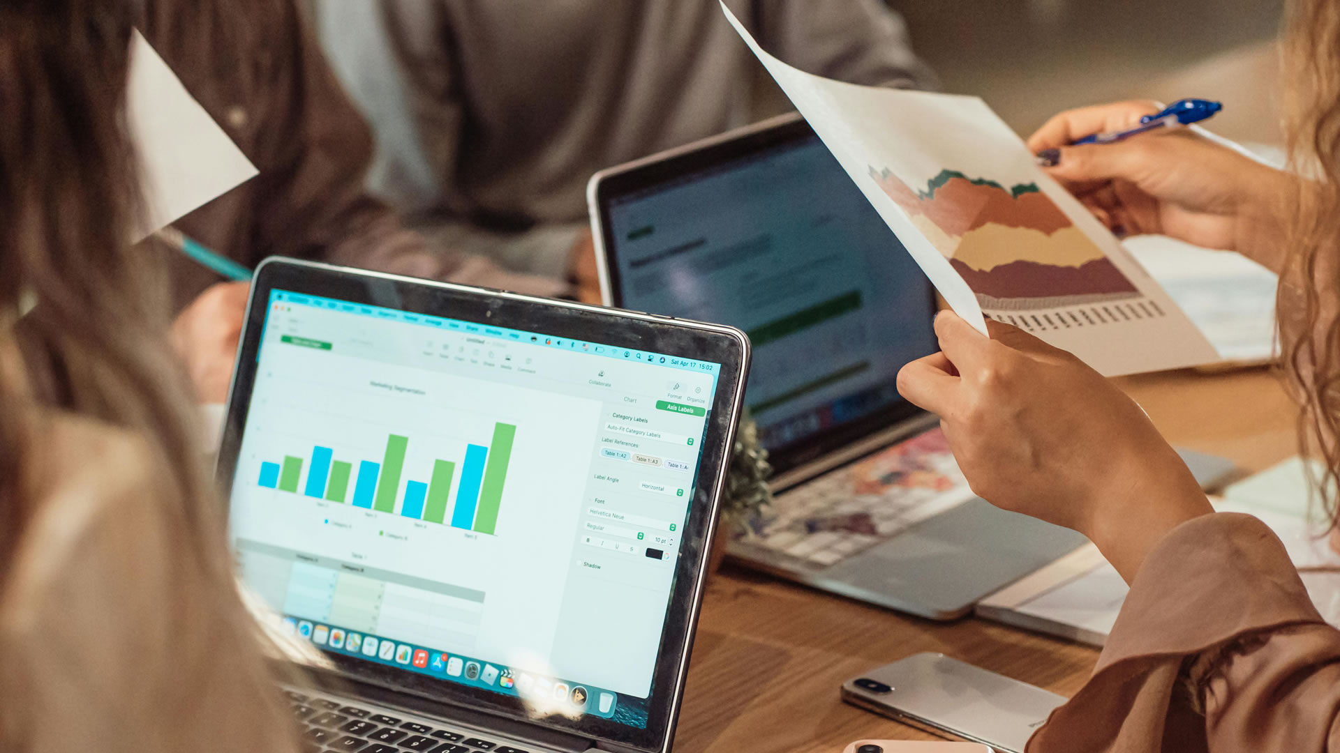 A group of people collaborating at a table, focused on laptops displaying various graphs and data analysis.
