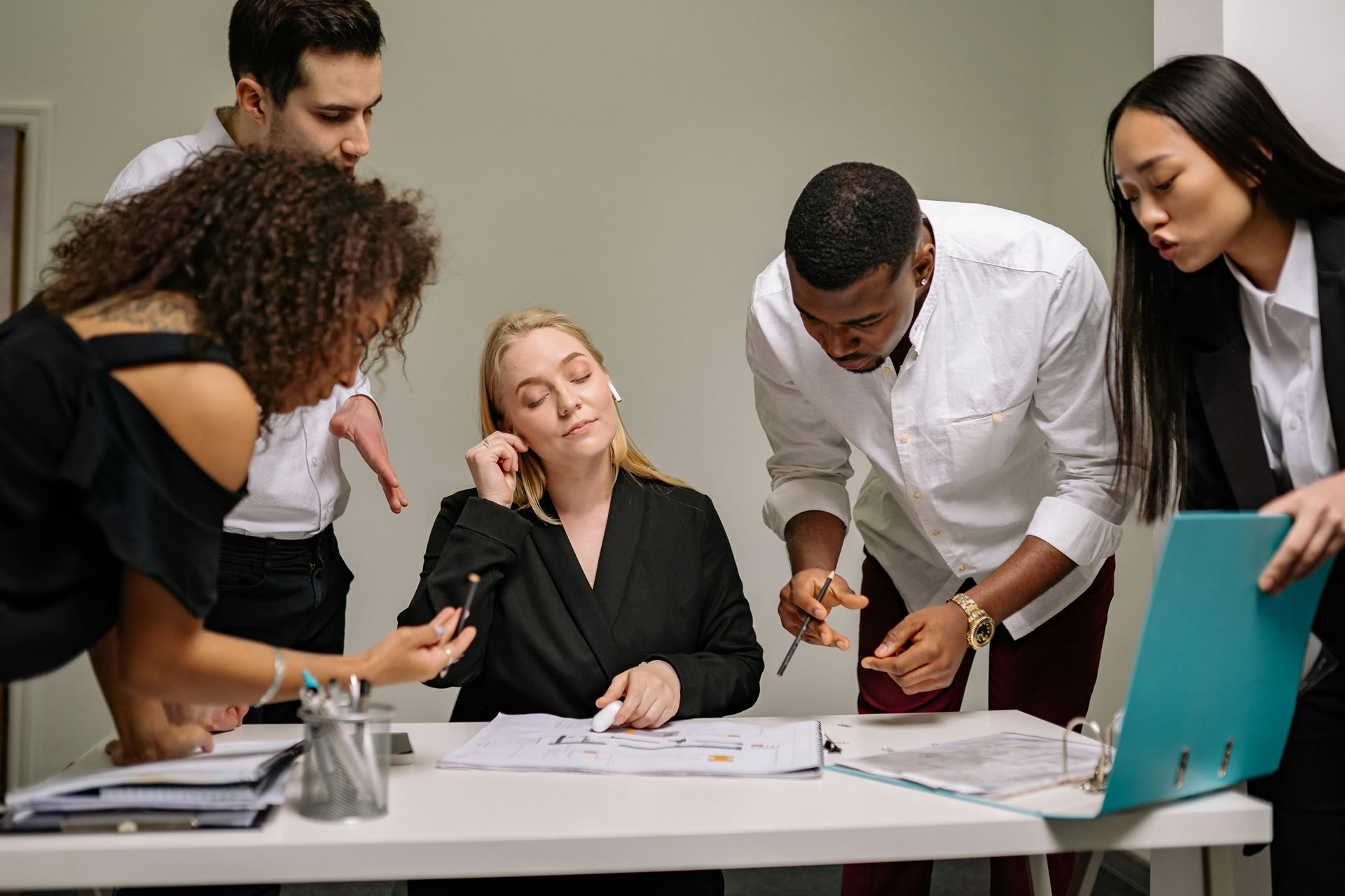 Overwhelmed businesswoman sitting at a desk with colleagues crowding around, symbolizing workplace stress and inefficiency.