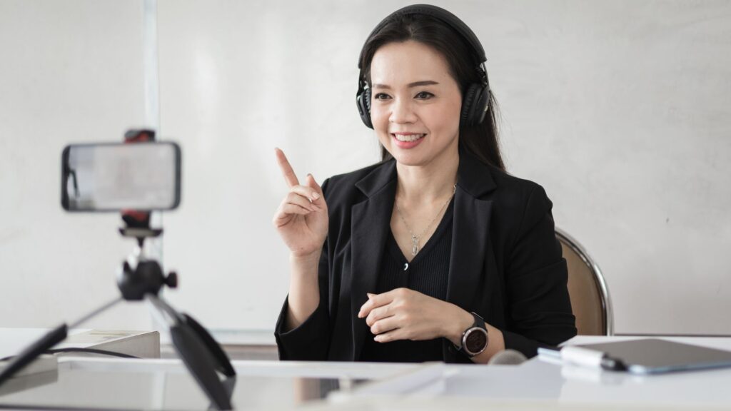 A professional woman wearing headphones, sitting at a desk and confidently recording a video using her smartphone on a small tripod.