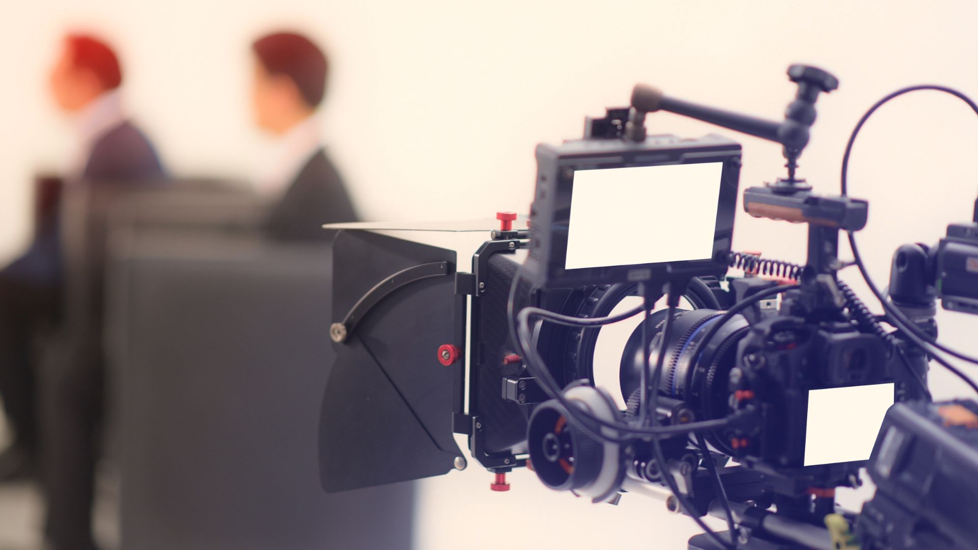 A close-up of a professional video camera with a monitor and lens hood, set up for a corporate video shoot with two people in suits blurred in the background.